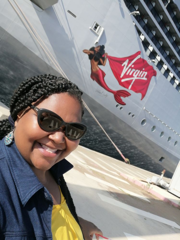 Jenita Lawal smiling in sunglasses, standing in front of the Valiant Lady's hull and mermaid logo
